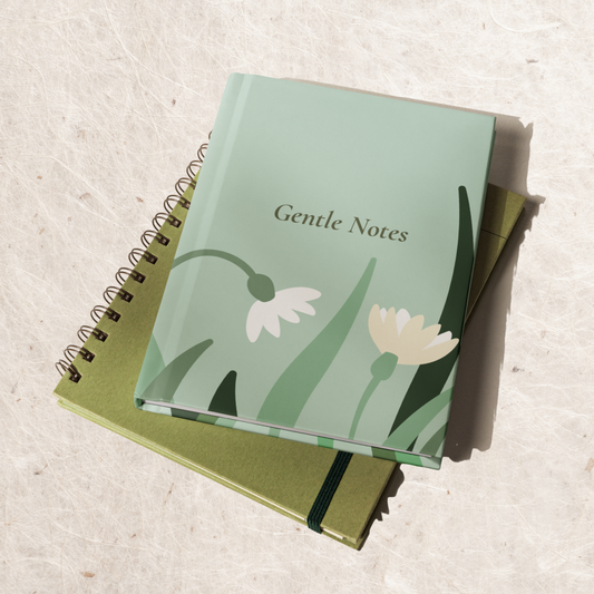 Sage green ‘Gentle Notes’ journal with white floral design, styled on top of two notebooks on a light textured surface with soft natural sunlight.